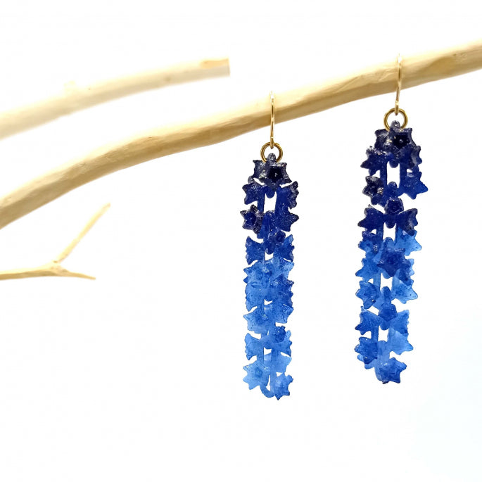 A pair of Blue Sakura Earrings from Tochu-De, shaped like cascading flowers, hang from a light-colored, textured branch against a white background. These floral accessories transition gracefully from deep blue at the top to a lighter blue at the bottom.