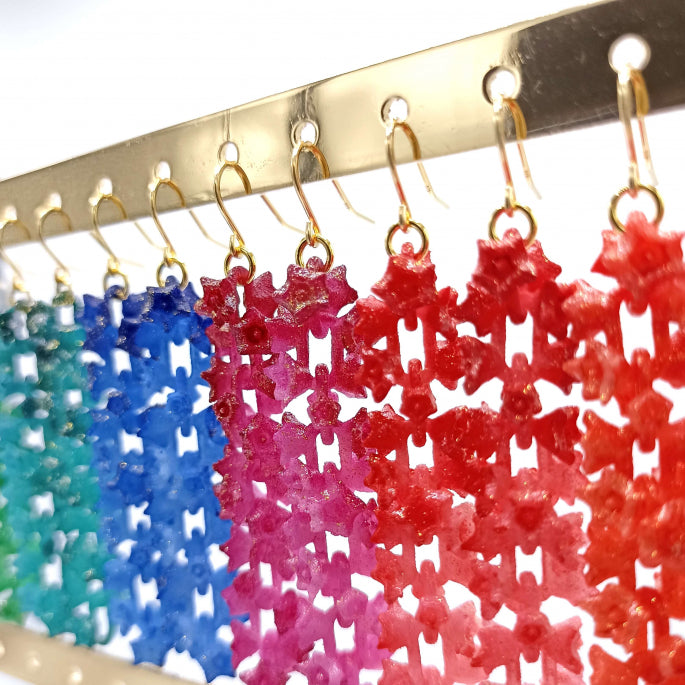 A close-up image of colorful, dangling earrings hanging on a gold display. The Tochu-De Fuchsia Sakura Earrings, part of the SAKURA Flower Jewelry collection, are intricately designed in styles resembling snowflakes or flowers, transitioning in color from green to blue to red. The white background highlights the vibrant colors.