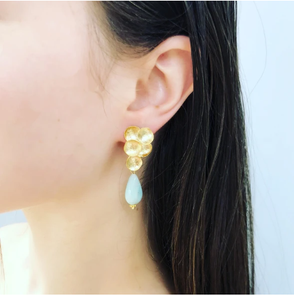 Close-up of a woman wearing the Aria V Bouquet Earrings, which are matte gold plated earrings shaped like a flower with polished amazonite teardrops hanging beneath them. Her dark hair is tucked behind her ear, and the background is a plain light color.