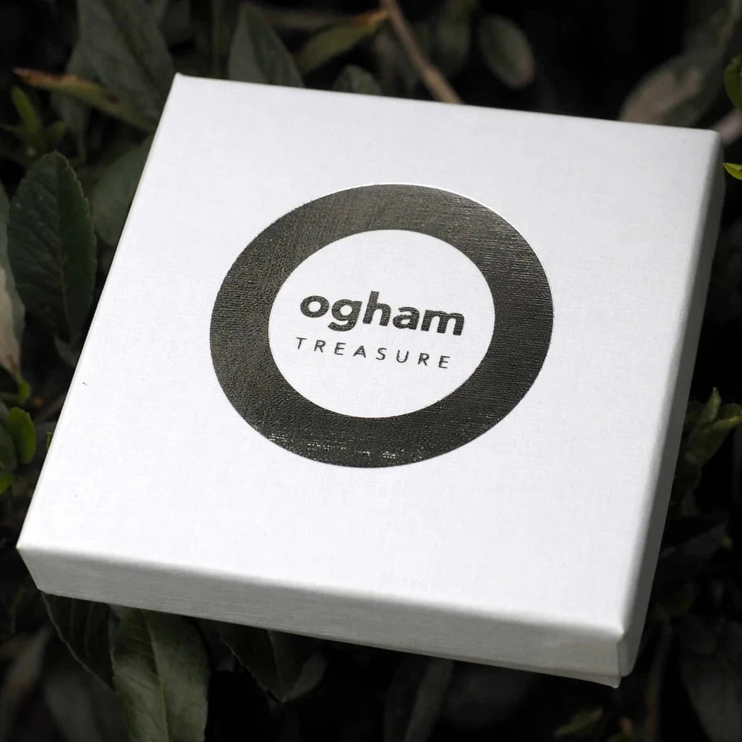 A white square box with the words "Ogham Treasure" in the center, surrounded by a thin black circle, rests among green leaves. Featuring an Ogham script and holding a sterling silver Love (Grá) Unisex Pendant, this elegant package captures ancient tradition and modern elegance beautifully.