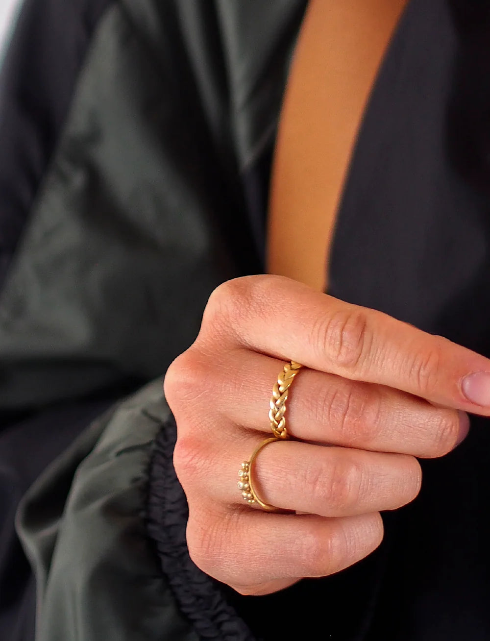A close-up of a hand wearing two gold rings, including Jane Koenig's Medium Braided Ring and another with small round details, set against a dark jacket sleeve.