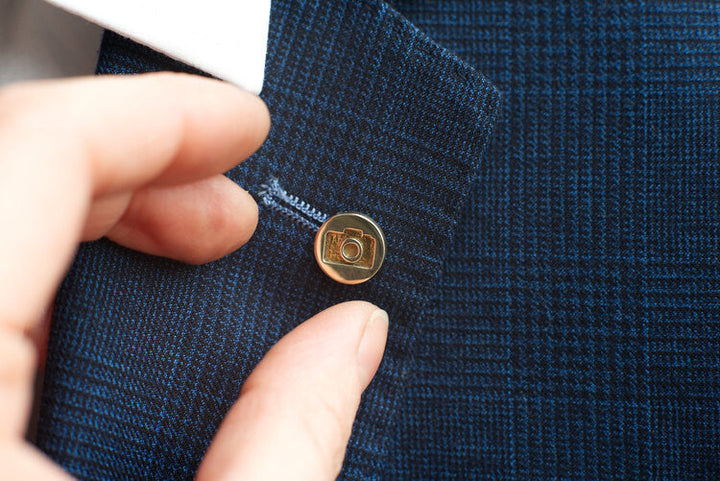 Close-up of a hand fastening a brass-colored button on a navy blue, plaid-patterned garment. The button, resembling the Millett Wade Camera Lapel Pin, features a small square camera icon engraved on it.