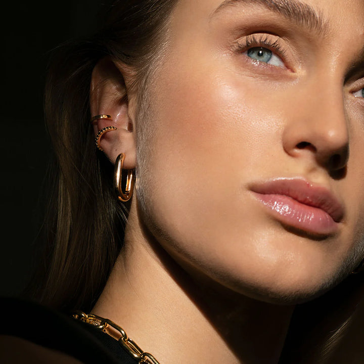 Close-up of a woman with light skin wearing Sif Jacobs Capri Medio Pianura Oval Hoop Earrings, a gold chain necklace, and several gold ear cuffs. She looks slightly upward with natural makeup and smooth, glowing skin.