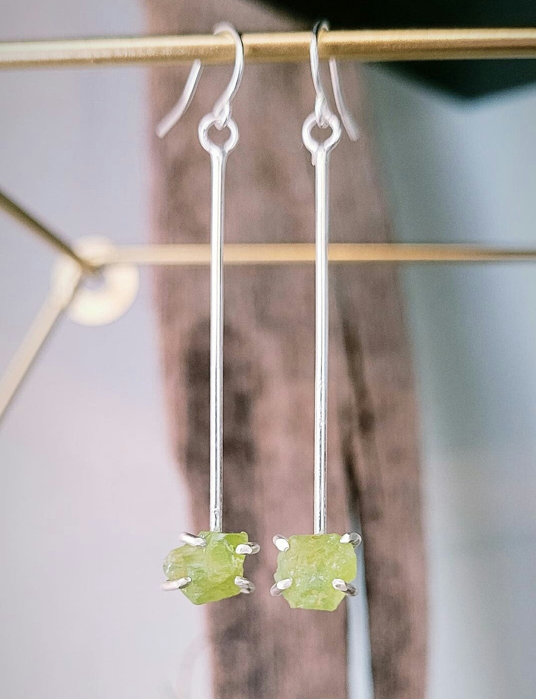 A pair of Raw Peridot Drop Earrings featuring long, slender sterling silver bars with raw Peridot gemstone accents at the ends. The earrings are displayed hanging from ear wires, with a blurred background that includes hints of a geometric shape and soft brown fabric. These exquisite pieces are crafted by Caroline Stokesberry-lee.