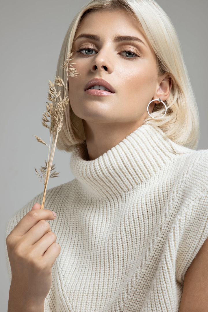 A person with short, blonde hair is wearing a white, chunky-knit turtleneck sweater. They are holding a dried plant close to their face and have a neutral expression. Accentuating their look are Carran Hoops in Sterling silver by Lynsey de Burca. The background is plain and light-colored.