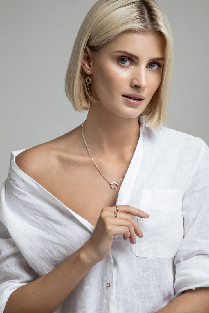 A person with short, blonde hair is wearing a white shirt slightly off one shoulder. They have a Sterling silver pendant, matching Carran earrings, and a Carran Mix Pendant by Lynsey de Burca. The background is plain and light-colored. The person is looking directly at the camera with a thoughtful expression.