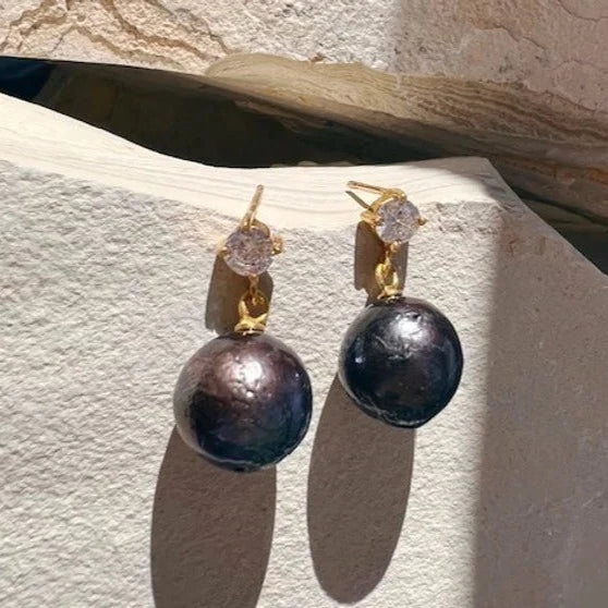 The Cassandra Earrings by Shyla, shown in various colours, feature gold on sterling silver with round black freshwater pearls and clear stone studs, displayed elegantly on a light stone surface in natural light.