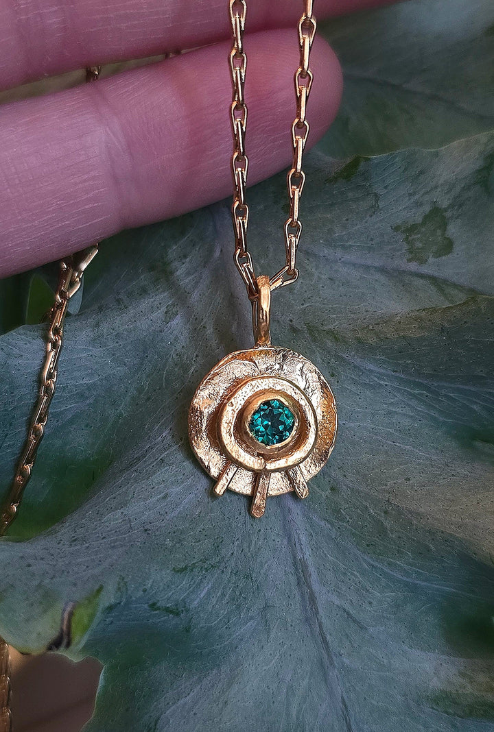 The Caroline Stokesberry-lee Celestial Eye Pendant Gold in Emerald showcases a textured gold-plated sterling silver circle with an emerald centerpiece, displayed against a large green leaf and partially held by a hand at the top.