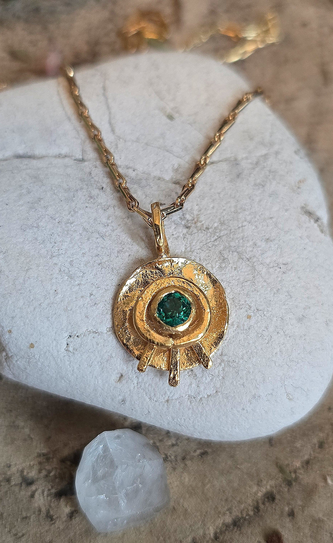 The Celestial Eye Pendant Gold in Emerald by Caroline Stokesberry-lee features a textured, gold-plated sterling silver circular design with an emerald green centerpiece, shown on a white stone beside a clear crystal.