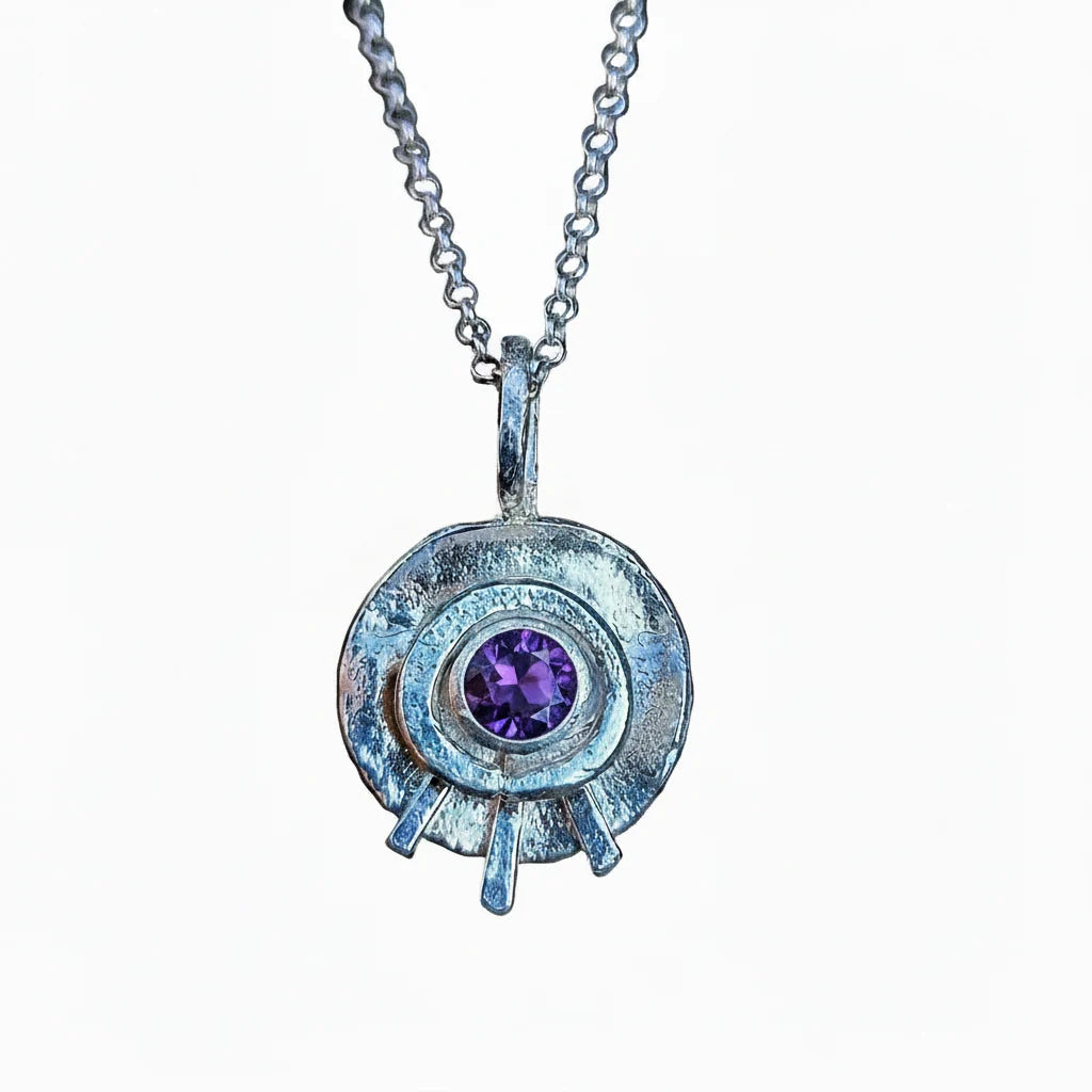 A silver pendant necklace featuring a textured circular disc with a central round purple gemstone, suspended from a silver chain against a plain white background.