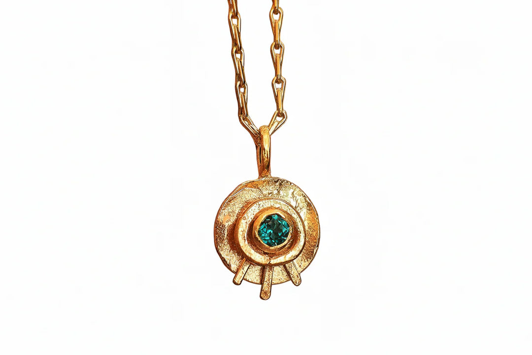 The Celestial Eye Pendant Gold in Emerald by Caroline Stokesberry-lee is a gold plated sterling silver necklace with a circular textured design and an emerald gemstone at the center, hanging from a gold chain.