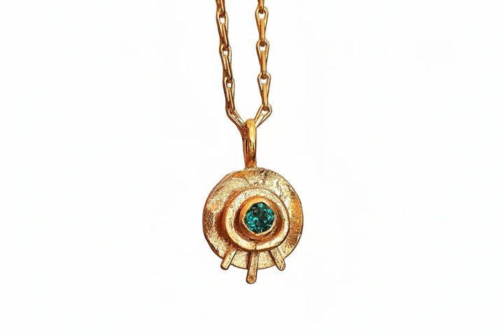 The Celestial Eye Pendant Gold in Emerald by Caroline Stokesberry-lee is a gold plated sterling silver necklace with a circular textured design and an emerald gemstone at the center, hanging from a gold chain.