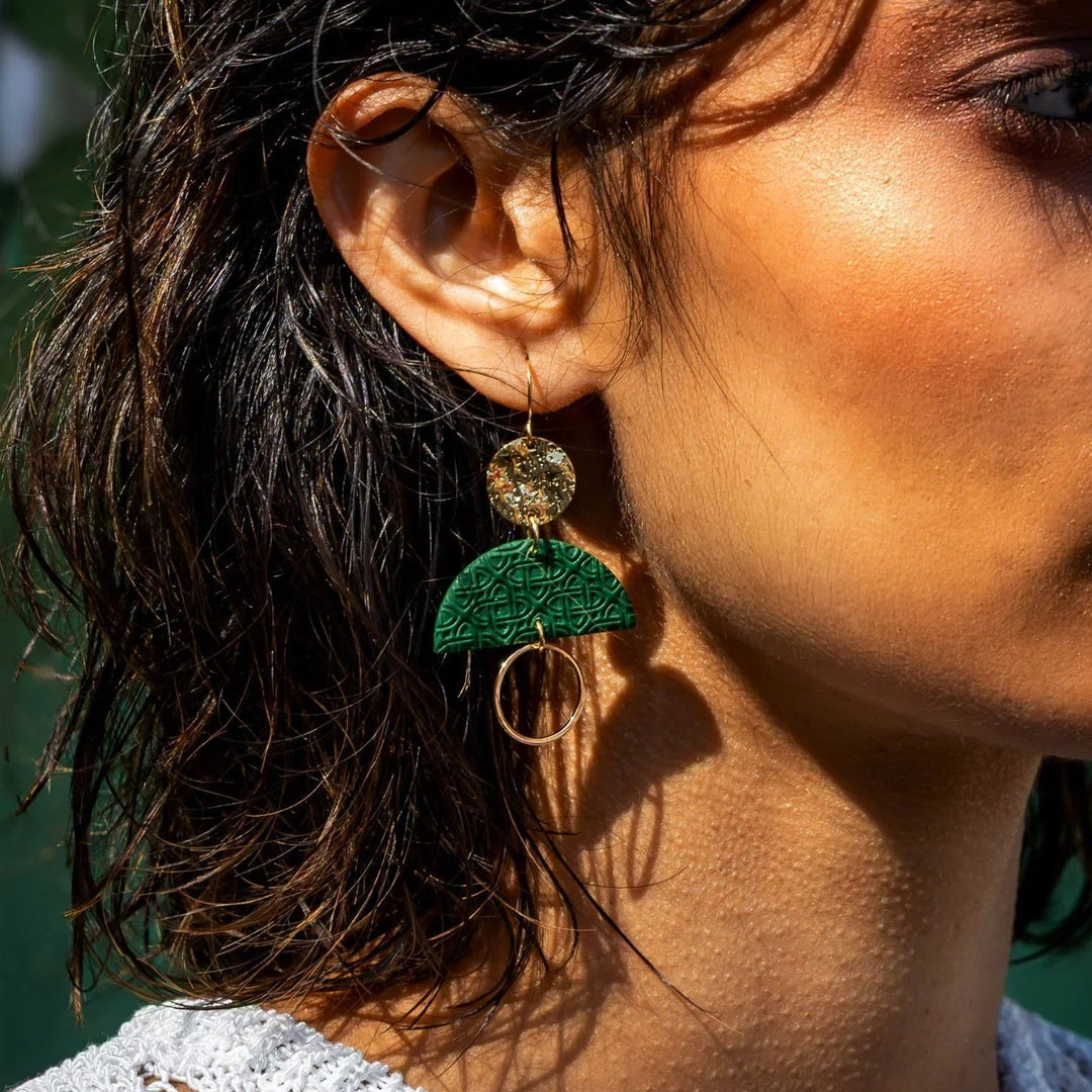 Close-up of a woman wearing a green leaf-shaped earring with a textured gold circle, against a blurred natural background.