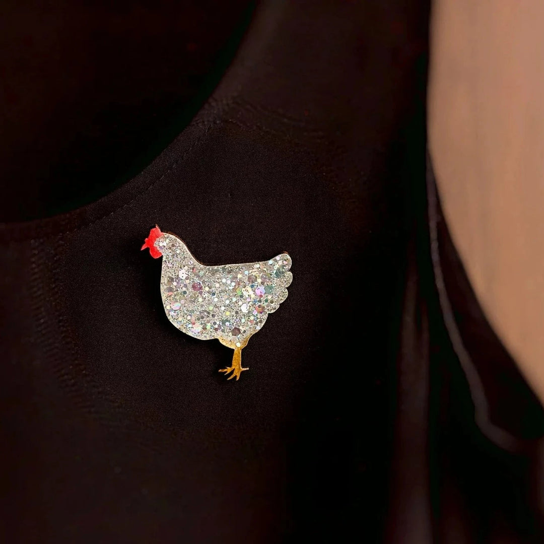 A close-up image of a person wearing a black garment with a glittering chicken-shaped brooch pinned on it. The Naoi Chicken Pin Brooch Silver Medium, reminiscent of handpainted wooden chickens, features a silver, sparkly body, a red comb, and a yellow leg, adding a whimsical touch to the outfit.