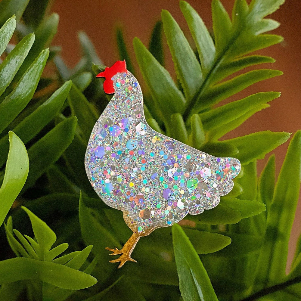 A glittery, holographic Chicken Pin Brooch Silver Medium by Naoi is placed among vibrant green leaves, sparkling with shades of blue, purple, and silver. It features a small red comb and yellow legs, evoking the charm of handpainted wooden chickens.