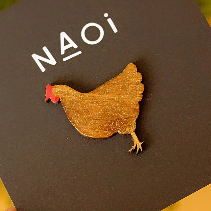 A Chicken Pin Brooch Gold with a red comb and wattle is pinned to a black card with the brand name "Naoi" printed in white letters at the top.