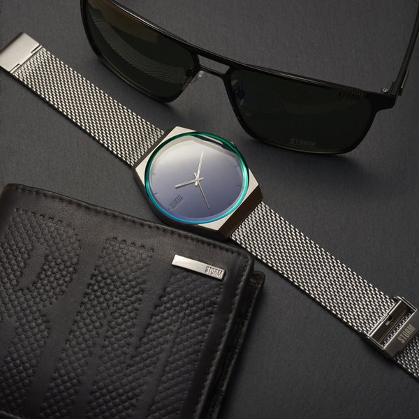 A **Storm Cirero Blue Watch** with a silver mesh strap and a blue gradient face is laid on a dark surface next to a pair of black sunglasses and a black leather wallet with a textured "STORM Cirero" design, adding a contemporary feel.