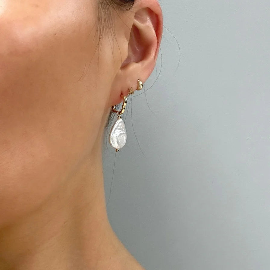 Close-up of a person’s ear wearing several earrings, including Aria V Clover Pearl Earrings with large, irregular-shaped freshwater pearls, and small gold studs, set against a plain light gray background.