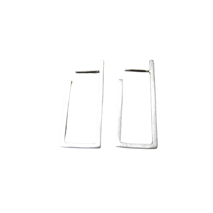 A pair of sleek, rectangular-shaped silver earrings from the MarCucurella Cub collection are placed against a plain white background. These handmade jewelry pieces feature minimalist, architectural lines with a rectangular frame and a small hoop closure at the top.