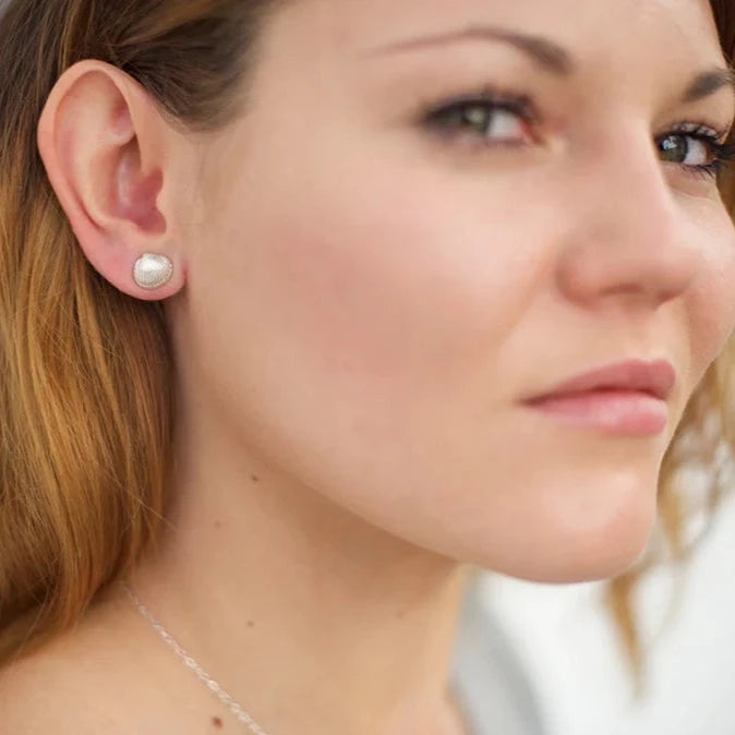 A close-up photograph of a woman with light brown hair. She is wearing **Tiny Cockle Shell Stud Earrings** by **Jennifer Kinnear** and has a subtle, gold plated silver necklace around her neck. The background is blurred, drawing focus to her facial expression and accessories.