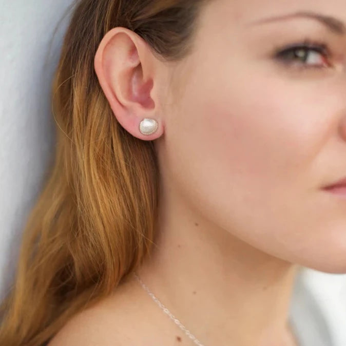 A close-up image of a person with long, light brown hair, wearing Jennifer Kinnear Tiny Cockle Shell Stud Earrings and a delicate silver necklace. The person's face is partially visible, looking slightly away from the camera.