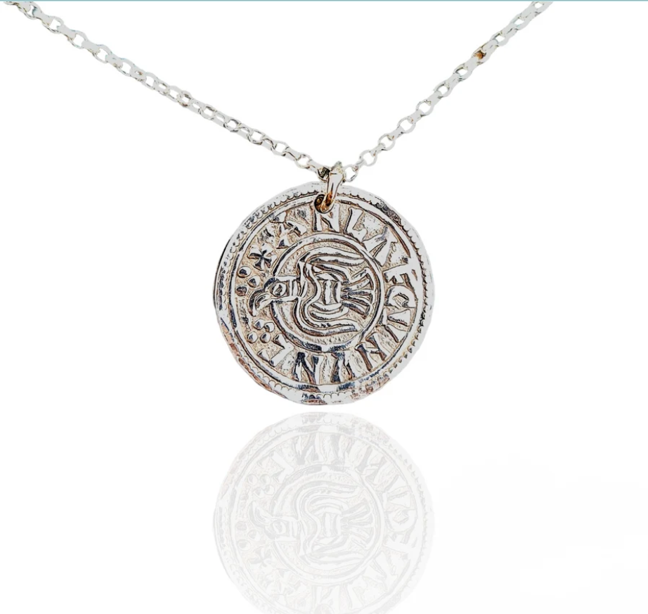 A silver chain necklace featuring a circular coin-style pendant with embossing, shown on a clean white background with a soft reflection below.