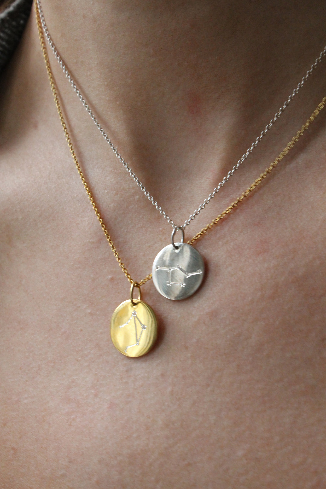 Close-up of a person's neck wearing two necklaces: the Symbol Necklace in 9k Gold by Personalised Charms and a silver necklace, each with a round constellation-engraved pendant.