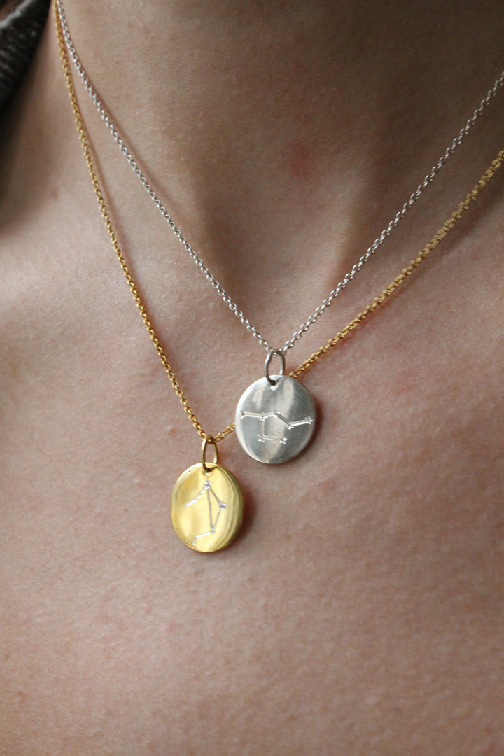 Close-up of a person's neck wearing two necklaces: the Symbol Necklace in 9k Gold by Personalised Charms and a silver necklace, each with a round constellation-engraved pendant.