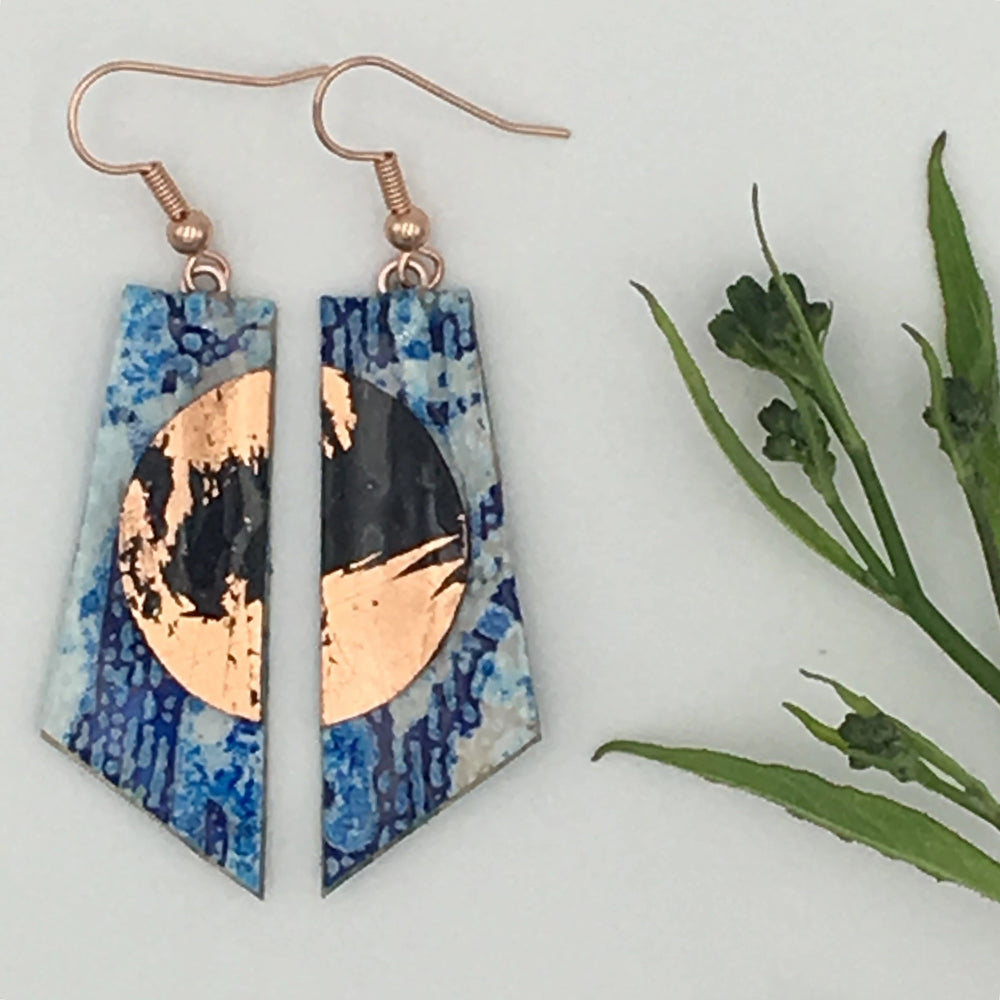 A pair of Cora Batik Textile Earrings in Blue/Black/Rose Gold from Rothlú with a geometric design, featuring a copper circular accent at the center and decorated with a unique blue texture. The hypo-allergenic rose gold plated hooks add elegance, and the earrings are displayed next to green foliage on a white background.