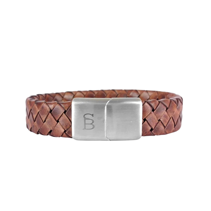 A braided brown genuine leather bracelet features a rectangular silver clasp engraved with the letters "SB," set against a white background. The **Preston - Caramel** by **Steel and Barnett** has a rustic yet refined appearance, suitable for both casual and formal occasions.