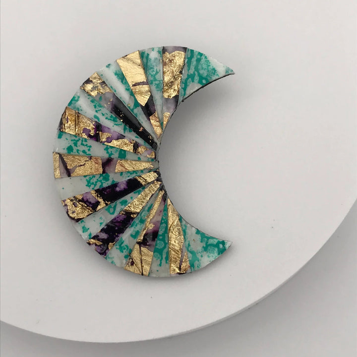 The Rothlú Crescent Textile Brooch in Mint/Gold/Aubergine showcases a mosaic pattern featuring gold leaf, green, purple, and white marble textures. Its intricate design includes geometric shapes radiating from the inner curve of the crescent and is enhanced by an eco-resin coating for added durability and shine. This striking and colorful piece of jewelry is truly unique.