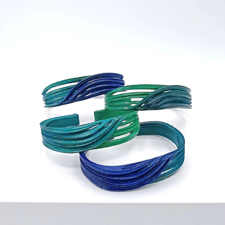 The Croisee Petrol & Emerald Bracelets by Tochu-De are lightweight, 3D printed jewelry pieces in blue and green with wavy openwork designs, made from bio-sourced materials and displayed upright on a white background.