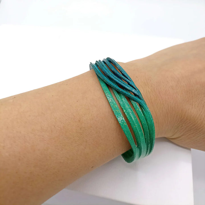A hand displays the Tochu-De Croisee Petrol & Emerald Bracelet, a lightweight, multi-strand leather accessory in green and blue tones, set against a white background.