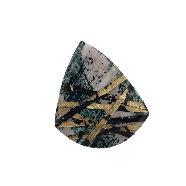 A polished, irregularly shaped gemstone with a mix of green, black, and beige colors. Gold-colored geometric patterns run across its surface like a batik tjanting technique, creating an intricate and modern design. The backdrop is plain black, highlighting the Rothlú Crosstown Ring Dark Green/Black/Gold's details.