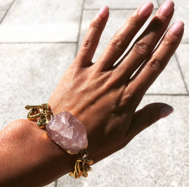 A hand wearing a Cruise Chunky Bracelet by Aria V with a large, rough-cut rose quartz stone is shown against a tiled pavement background. The fingernails are painted in a light pink shade. This exquisite piece, handmade in Dublin, radiates elegance and charm.