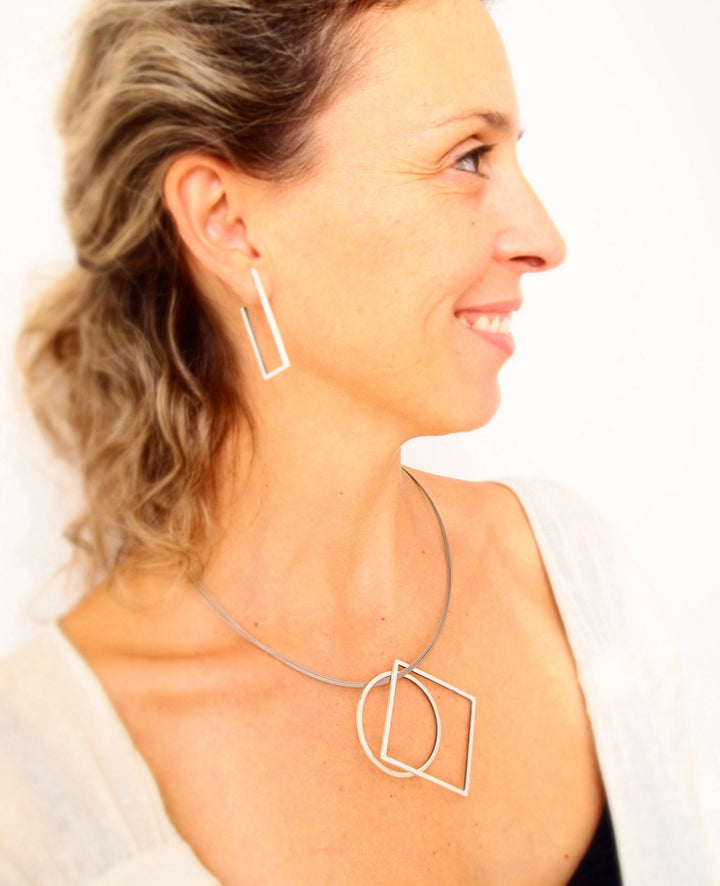 A woman with light hair and a light complexion, facing right and smiling. She is wearing handmade jewelry with architectural lines, including rectangular earrings and a MarCucurella Cub Pendant featuring matt silver square and circular pendants. She is dressed in a light-colored top against a white background.