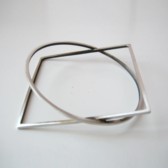 A modern minimalist silver bracelet, the Cub Silver Bracelet by MarCucurella, featuring an intertwined square and circular shape, inspired by architectural design, positioned on a plain white background.