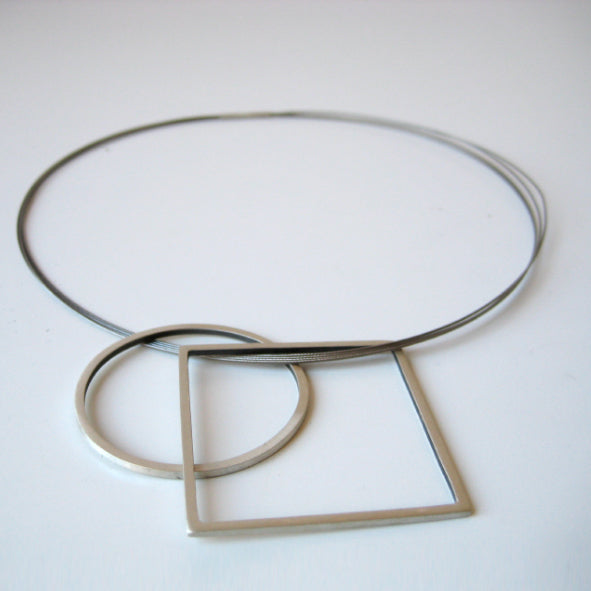 A minimalist necklace featuring a thin wire loop with two geometric pendants: one circular and one square. Handmade jewelry in matt silver, the Cub Pendant by MarCucurella is displayed on a white background, highlighting the architectural lines and simplicity of the design.