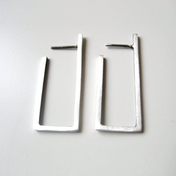 Part of the Cub collection, this pair of modern rectangular silver earrings showcases architectural lines and an open-ended geometric design with a brushed metal finish. Displayed on a plain white surface, each earring is handmade jewelry with the post conveniently positioned on top for easy fastening. These are the Cub Long Silver Earrings by MarCucurella.