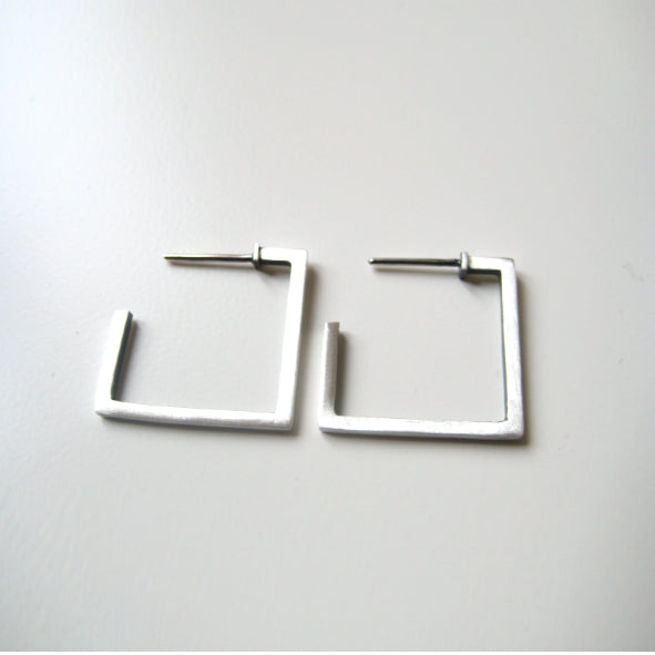 A pair of minimalist square-shaped silver earrings with post backs are placed on a clean, white surface. The handmade jewelry features geometric and modern design, showcasing sleek architectural lines in oxidized silver for a unique and contemporary look. These are the Cub Square Earrings by MarCucurella.