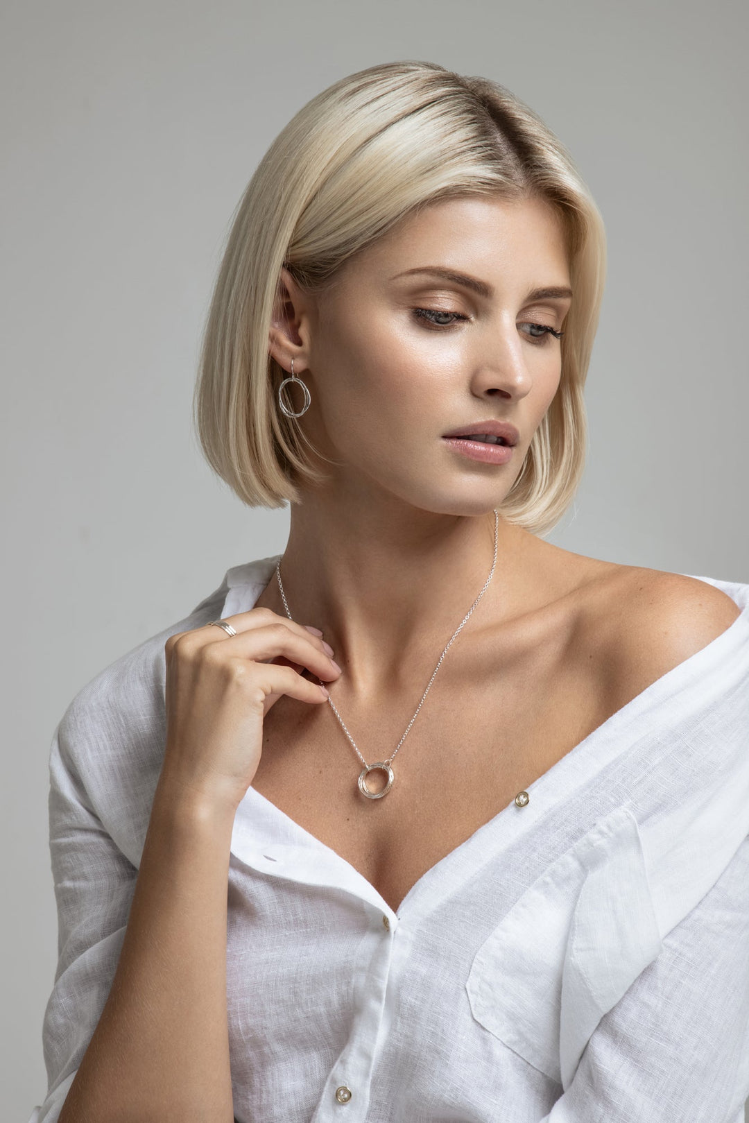 A woman with blonde shoulder-length hair wears a white shirt slightly off one shoulder, showcasing a Lynsey de Burca Doorus Mix Pendant and matching drop earrings. Both pieces are crafted in sterling silver. She has a serene expression and looks away from the camera, gently touching her shirt's collar.