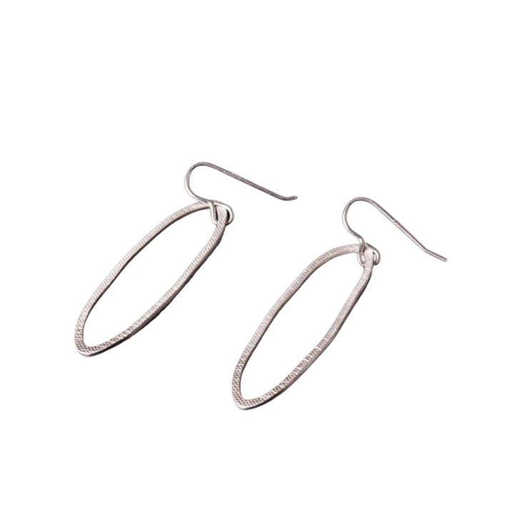 A pair of Lynsey de Burca Drift Drop Earrings with hook attachments, resting against a white background. The hand-forged earrings feature a sleek, elongated oval design with a slight texture along the edges, reminiscent of fishing net rope.