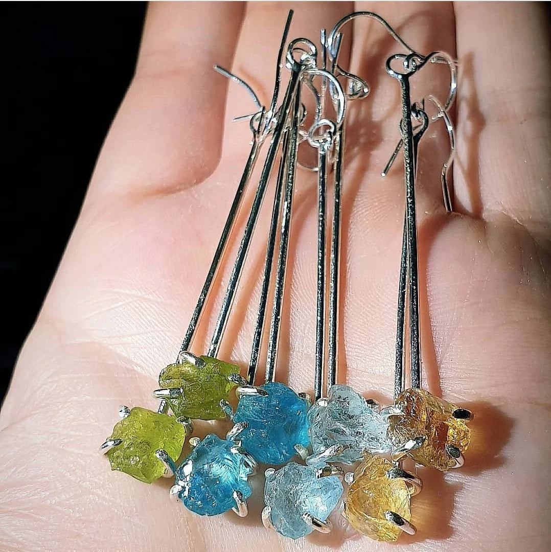 A hand is holding a collection of Raw Aquamarine Drop Earrings by Caroline Stokesberry-lee, each adorned with raw, unpolished gemstones in various colors: yellow, blue aquamarine gemstones, and light brown. The earrings, fashioned with sterling silver ear wires, are arranged side by side to showcase their distinct and natural textures.