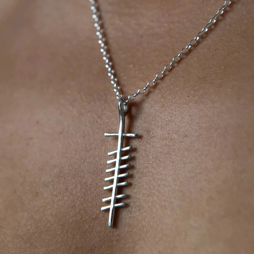 A close-up of a sterling silver fishbone pendant necklace worn on a person's neck. The Love (Grá) Unisex Pendant by Ogham Treasure features a minimalist design of a fish skeleton with a simple chain. Handcrafted in Ireland, the background is blurred, emphasizing the necklace.