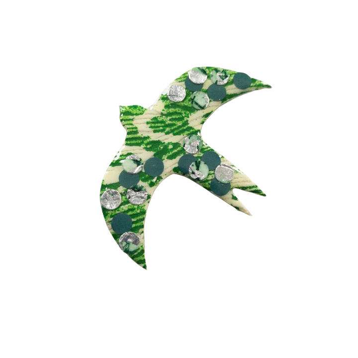 A decorative, green and white bird-shaped pendant with circular, metallic elements embedded on its surface, set against a plain, black background. The bird is in an abstract design with open wings and a pointed beak, showcasing a unique texture through an eco resin finish. This elegant piece is the Éan Bird Batik Textile Brooch by Rothlú.