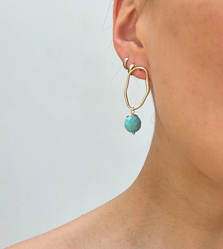 Close-up of a person's ear wearing Aria V's Eddie Earrings—modern gold-plated earrings with a large turquoise bead pendant. The plain background and partially visible shoulder highlight these striking turquoise earrings.