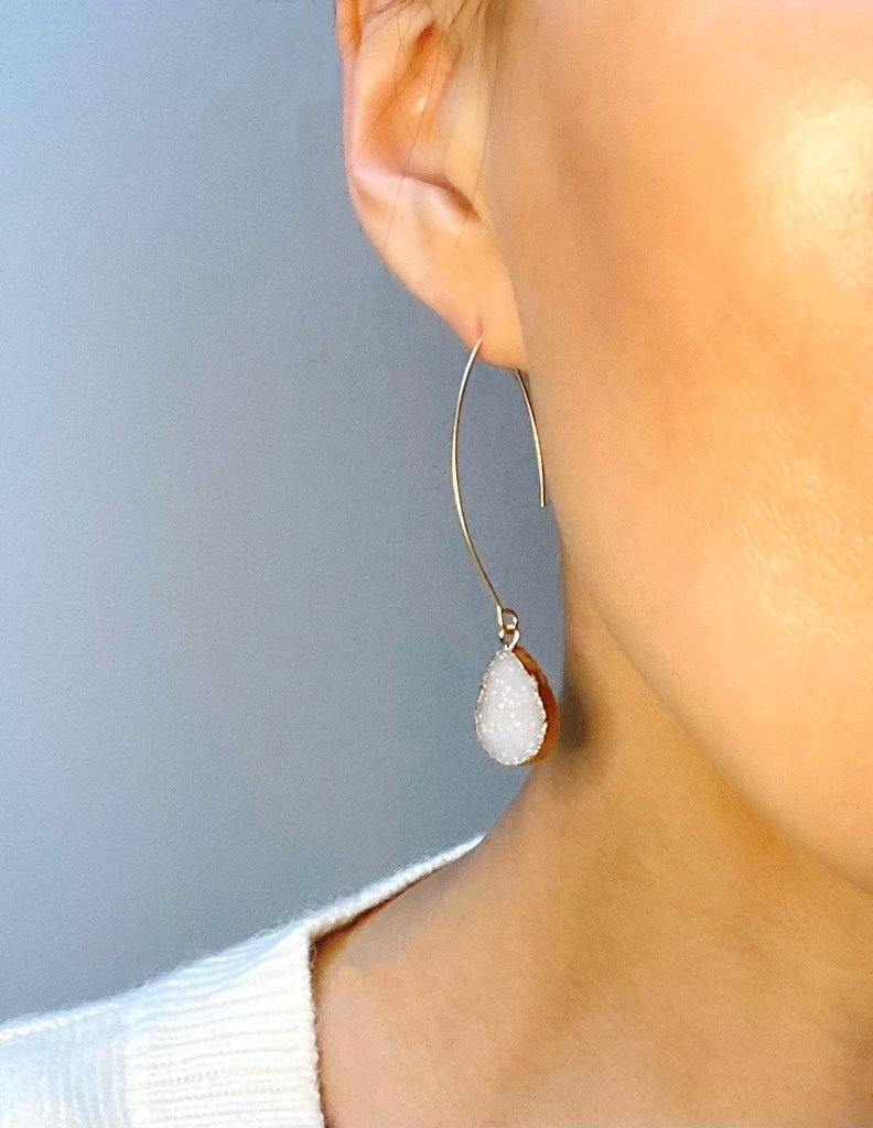 Close-up of a person wearing a handcrafted hoop earring with an AB white Druze quartz teardrop-shaped pendant. The person is facing slightly sideways, with the earlobe and cheek partially visible against a light background. They are wearing a light-colored top, showcasing the elegant Eira Earrings by Aria V.