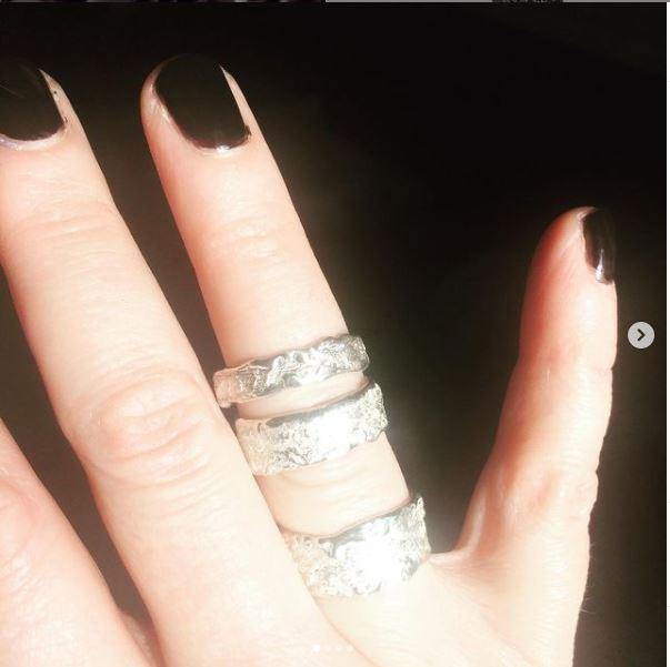 A hand with black nail polish is shown wearing three sterling silver rings (Elements Ring Wide by Abbie Dixon) on the middle finger, index finger, and thumb. The lighting emphasizes the details of each textured ring and creates a slight shadow on the hand.