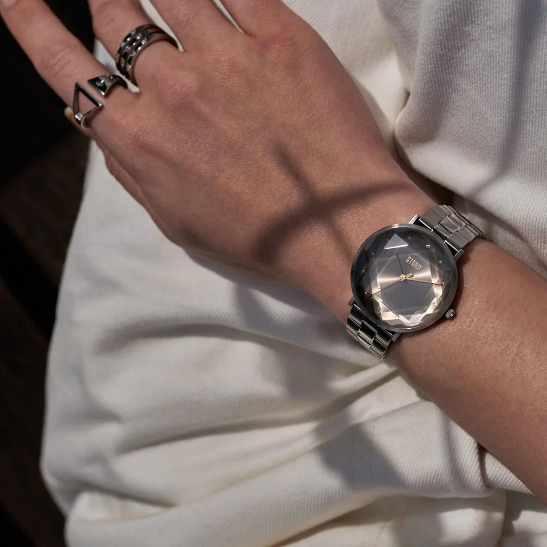 A person's left wrist adorned with a Storm Elexi Taupe watch by Storm Watches featuring a faceted circular face and two-tone stainless steel strap. The hand sports several rings, including a geometric triangle ring, while the individual is dressed in a long-sleeved white garment.