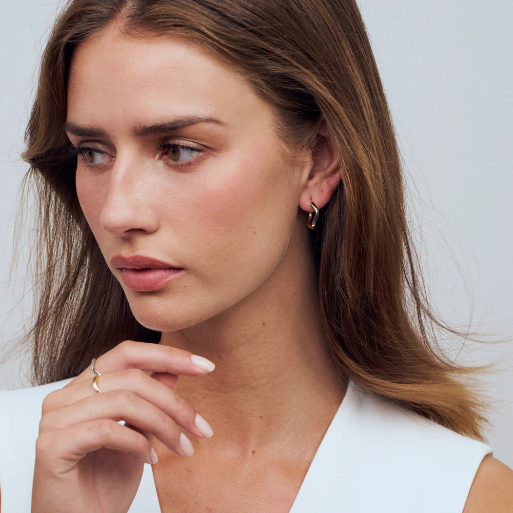 A woman with long brown hair, wearing a sleeveless white top and Sif Jacobs Ellera Waves Pianura Hoop Earrings, gazes thoughtfully to the side with her hand near her face, embodying timeless style.
